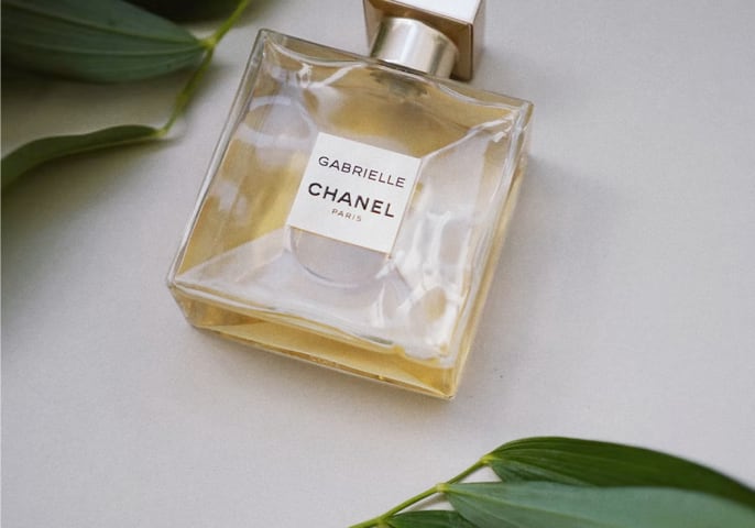 A gold-capped, clear glass bottle of Chanel Gabrielle perfume, resting on a white surface with green leaves.