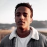 Young man with medium brown skin, 
              short dark hair, wearing a jacket with 
              a light-colored shearling collar, standing 
              outdoors with a blurred background and soft 
              evening light.